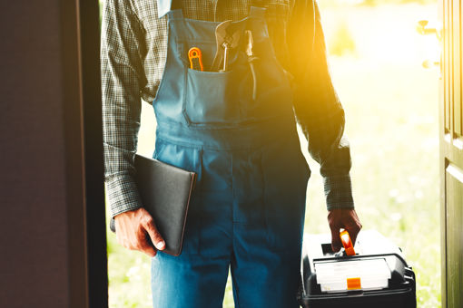 Contractor walking through front door wearing overalls and carrying a toolbox with sun shining in the background.